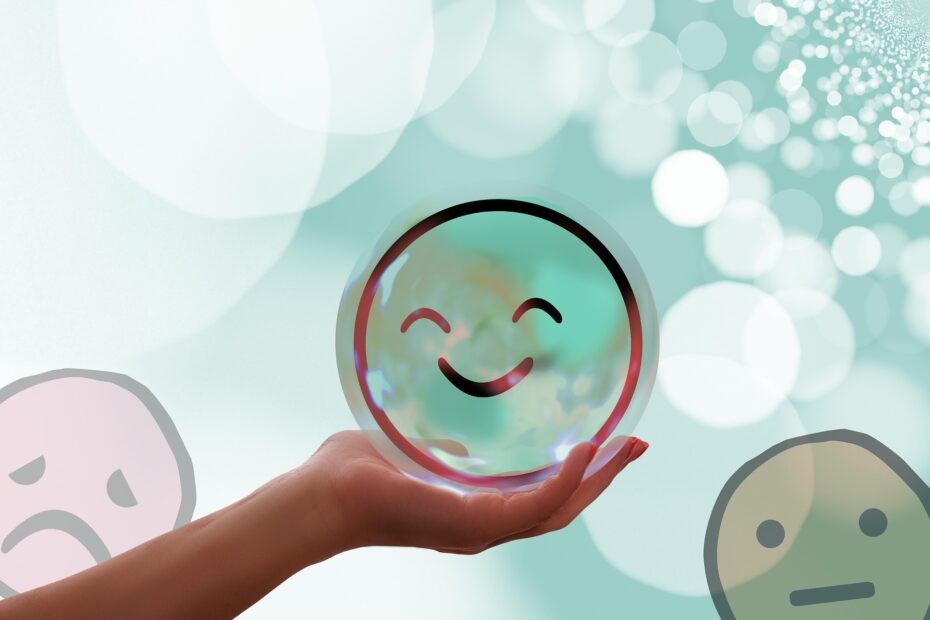 Hand, die einen lachenden Smiley hält - links im Hintergrund ein trauriger Smiley - rechts ein neutral blickender